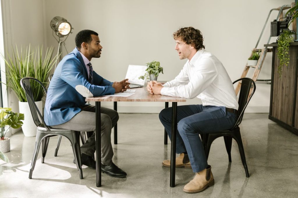 Two businessmen engaged in a professional discussion at an indoor office setting with greenery and modern decor.